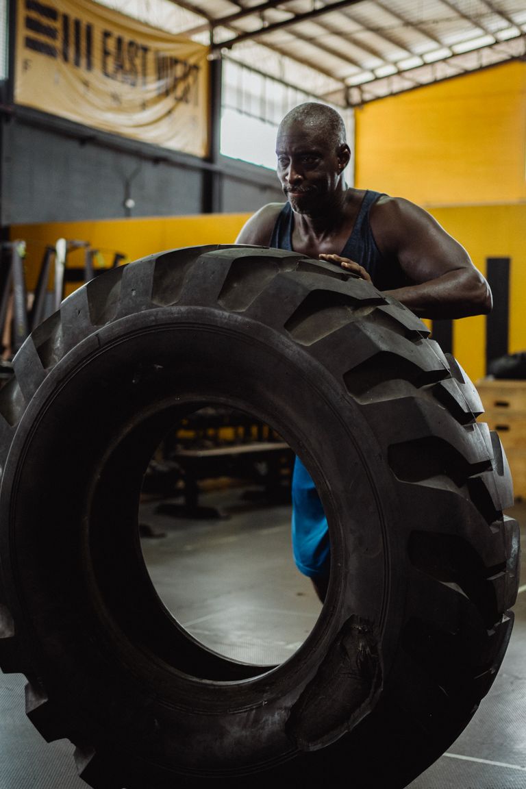 Athletischer Mann im blauen Tank-Top wuchtet einen schweren Traktorreifen in einer Industrie-Halle - hochintensives Krafttraining ab 40