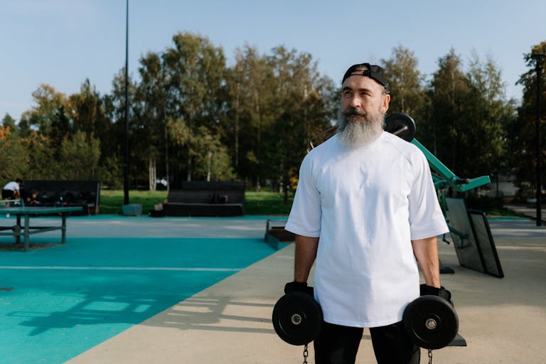 Mann mit weißem Bart und Cap steht mit zwei Kurzhanteln im herbstlichen Outdoor-Fitnesspark - Krafttraining wie 5x5 für Wiedereinsteiger ab 40