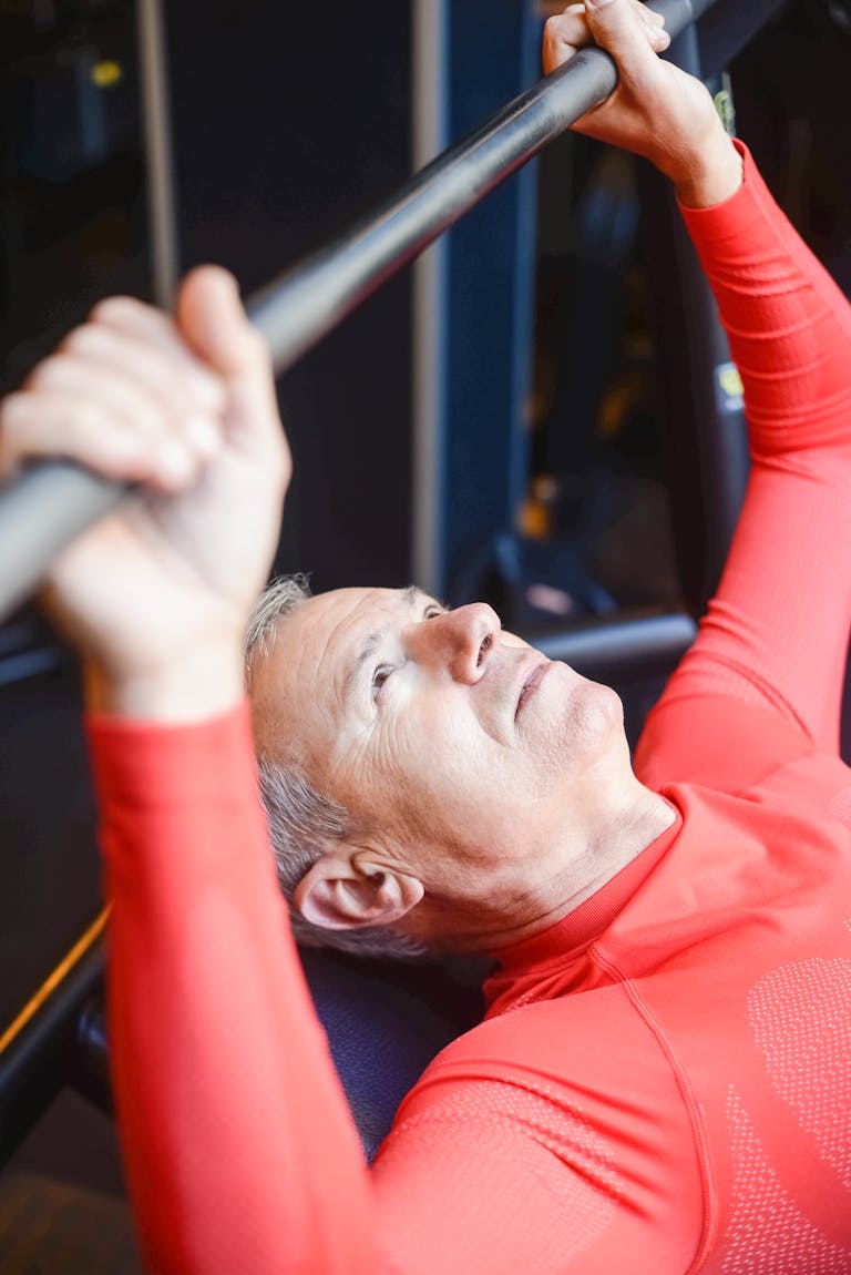 Mann ab 50 im roten Funktionsshirt drückt konzentriert eine Hantelstange auf der Bank im Fitnessstudio - gelenkschonendes Superslow-Training ab 40